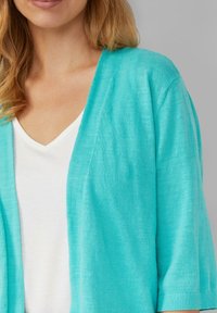 Cardigan turchese aperto davanti con maniche corte, texture leggera e vestibilità ampia. Indossato sopra una maglietta bianca a scollo a V.