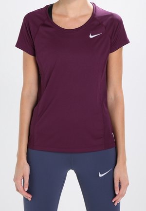 Frau trägt ein eng anliegendes weinrotes Nike-T-Shirt und dunkelblaue Leggings mit Nike-Logos auf schlichtem hellem Hintergrund.