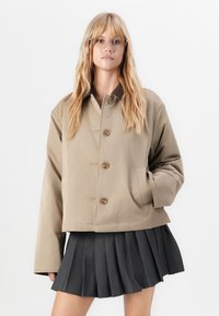Beige verkürzte Jacke mit großen Knöpfen, seitlichen Taschen und einem braunen Kragen. Kombiniert mit einem gefalteten schwarzen Minirock. Glatte Textur.