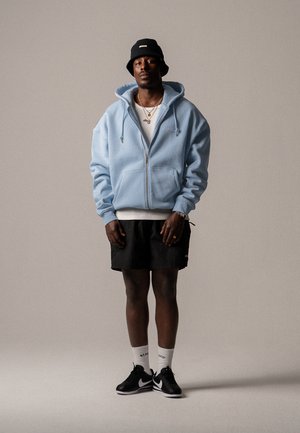 Un sweat à capuche zippé bleu clair avec cordons, un short noir, des baskets Nike noires et un chapeau seau bleu marine ; un t-shirt blanc et des bijoux accentués.