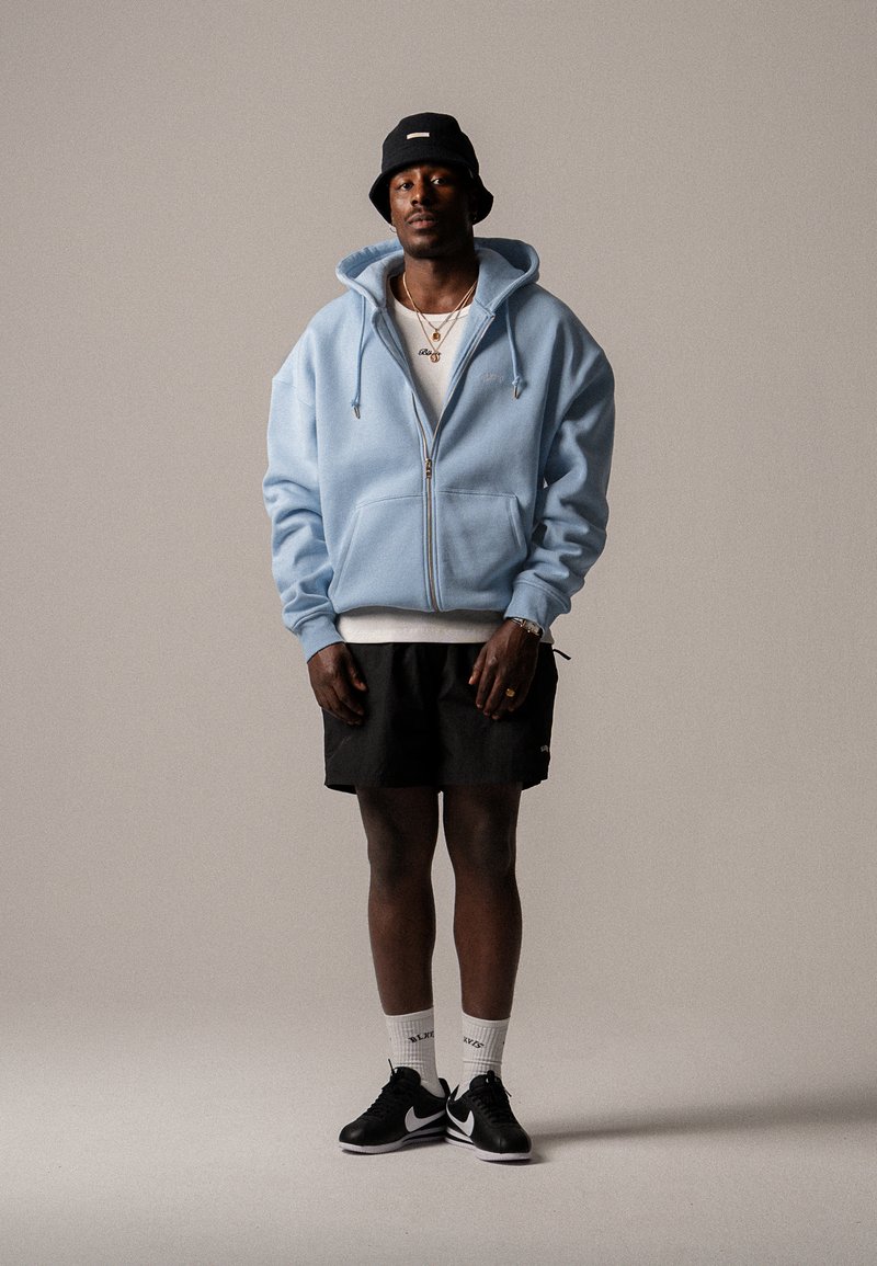Hellblaue Reißverschlussjacke mit Kordelzügen, schwarze Shorts, schwarze Nike-Sneaker und ein navy-blauer Bucket Hat; weißes T-Shirt und auffälliger Schmuck.