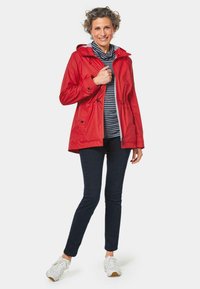 Rote wasserdichte Jacke mit Kapuze, ausgestattet mit Kordelzügen und einem Reißverschluss, kombiniert mit einem gestreiften Hemd, dunklen Hosen und weißen Turnschuhen.