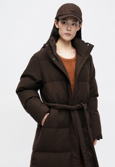 Lang brun dunjakke med bælte i taljen, lynlåslukning og høj krave. Båret over en orange sweater, parret med en brun kasket.