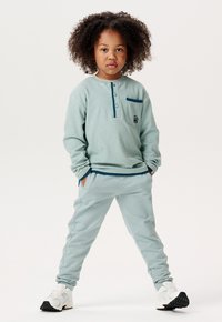 Helles blaues Langarmhemd mit Henley-Kragen und einer kleinen Tasche; passende hellblaue Hose; weiße und schwarze Sneakers.