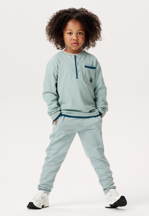 Helles blaues Langarmhemd mit Henley-Kragen und einer kleinen Tasche; passende hellblaue Hose; weiße und schwarze Sneakers.
