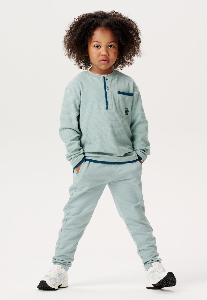 Helles blaues Langarmhemd mit Henley-Kragen und einer kleinen Tasche; passende hellblaue Hose; weiße und schwarze Sneakers.