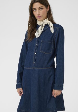 Femeie purtând o rochie din denim bleumarin, cu buzunar frontal, nasturi pe față, mâneci lungi și un fular alb cu model la gât.
