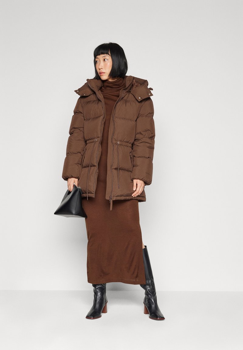 Brązowa kurtka puchowa z kapturem, z wciągnięciem w pasie, zapinanymi kieszeniami, noszona na brązowej sukience z golfem i połączona z czarnymi botkami na obcasie.