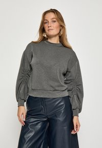 Sudadera gris de cuello redondo con mangas abullonadas y detalle de fruncidos en los laterales, combinada con pantalones de cuero azul marino de cintura alta. Textura suave, ajuste casual.