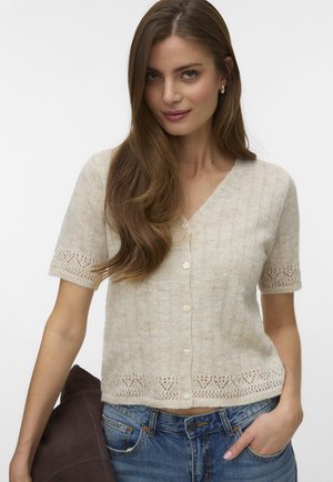 Frau mit langen braunen Haaren, die ein beiges kurzärmeliges Strickoberteil mit Knöpfen und blaue Jeans trägt und in einer Hand eine dunkle Jacke hält.