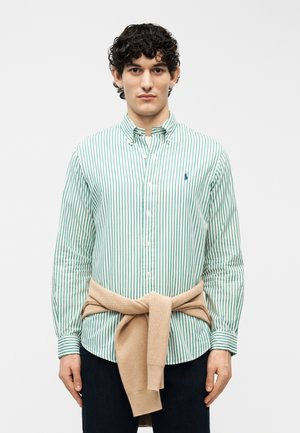 Homme aux cheveux bouclés portant une chemise boutonnée à rayures verticales vertes et blanches, avec un pull beige noué autour de la taille, sur un fond uni.