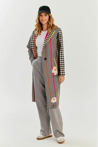 Długi płaszcz w kratkę houndstooth z dużymi haftami kwiatowymi, kolorowymi paskami, czarnymi guzikami i transparentnymi rękawami. Połączony z szarymi spodniami i trampkami.
