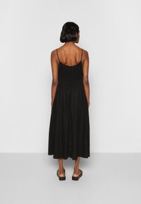 Vestido preto de comprimento médio com alças finas e cintura franzida, apresentando uma saia fluida. Usado com calçado plano, proporcionando um ajuste descontraído.