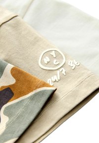 Vêtement en coton présentant une variété de couleurs : un motif camouflage beige, vert et orange. Comprend la marque "NYC" en blanc, avec des détails en surpiqûres.