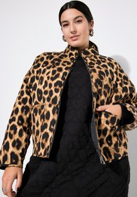 Giacca leopardata in marrone e nero, con colletto alto, zip frontale e tasche laterali. Indossata sopra un abito nero con texture.
