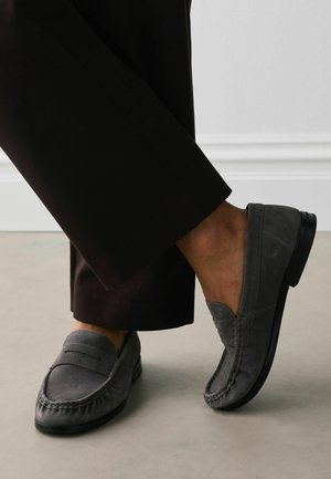 Person wearing dark gray suede loafers and black cropped trousers, standing on light gray floor near white wall baseboard.