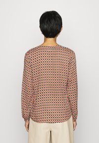 Blouse à manches longues en tissu à motifs, présentant un design géométrique de losanges oranges, verts et blancs sur un fond marron.