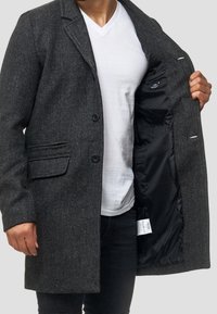 Abrigo gris texturizado con solapa con muesca, cierre de un solo botón y dos bolsillos delanteros. El interior es de satén negro con una etiqueta visible.