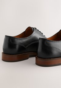 Schwarze Lederschuhe mit glatter Textur, brauner Ledersohle und minimalen Nähdetails an Ferse und Seiten. Sichtbares Schnürdesign.