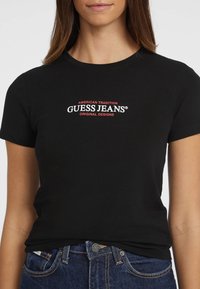 Čierne bavlnené tričko s okrúhlym výstrihom, s vyšívaným textom v červenej a bielej farbe "GUESS JEANS" a "AMERICAN TRADITION" nad ním.