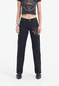 Black denim jeans with a loose fit, featuring embroidered snake and floral designs. Paired with a fitted black graphic t-shirt, cropped length.