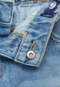 Tă fabrică de denim de culoare albastru deschis, arătând un prim-plan al unui buton și cusăturii. Prezintă un buton argintiu cu un centru auriu și o ață contrastantă de culoare bej.