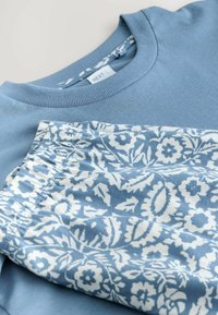 Kék rövid ujjú ing bordázott nyakkivágással, kék-fehér virágmintás, gumis derekú rövidnadrággal párosítva.