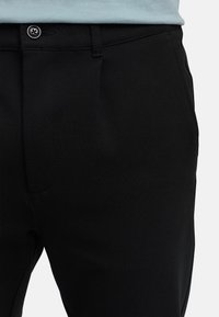 Nærbilde av svarte bukser med knappet linning og sidelommer, brukt med en lyseblå skjorte som er stukket inn.