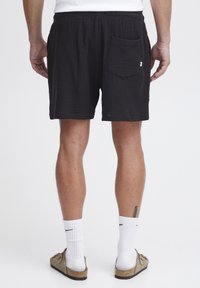 Shorts negros texturizados con cintura elástica y un bolsillo trasero. Usados con calcetas blancas y sandalias beiges. Vista desde atrás.