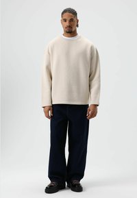 Sweat-shirt beige surdimensionné avec un col côtelé, associé à un jean bleu foncé à jambes larges et des chaussures à plateforme noires texturées.