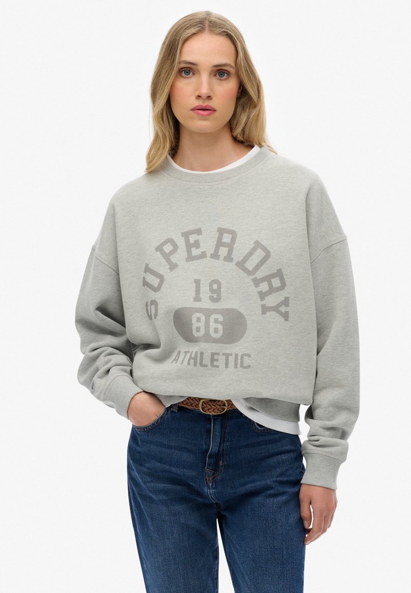 Sudadera gris corta con "SUPERDRY 1986 ATHLETIC" impreso en un gris más oscuro. Cuello redondo, corte relajado y puños acanalados. Combinada con jeans azules.