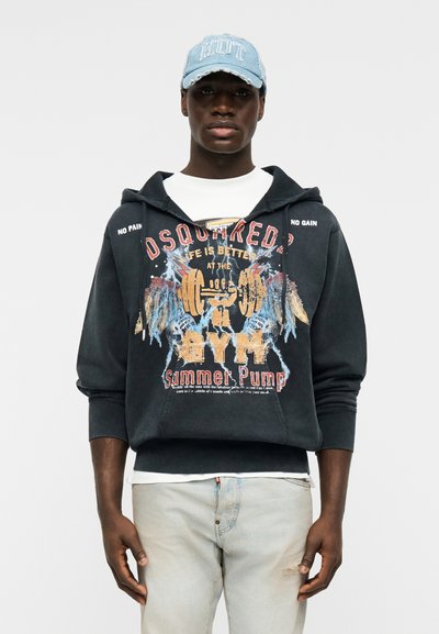 Homme portant un sweat à capuche noir avec un texte graphique sur le thème de la salle de sport et une casquette en denim bleu clair, associé à un jean délavé clair.