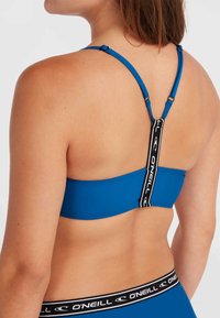 Top sportivo blu con spalline regolabili, caratterizzato da una fascia nera a contrasto con il nome del brand. Texture liscia, design minimalista.