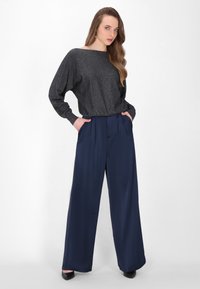 Zwarte glinsterende pullover met lange mouwen, gecombineerd met marineblauwe broek met wijde pijpen. Beide kledingstukken hebben zachte texturen en een op maat gemaakte pasvorm.