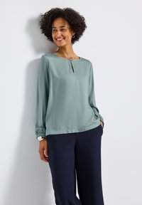 Blusa verde menta a maniche lunghe con scollatura rotonda e dettaglio a chiave. Presenta polsini arricciati e un tessuto leggero e liscio. Abbinata a pantaloni blu navy.