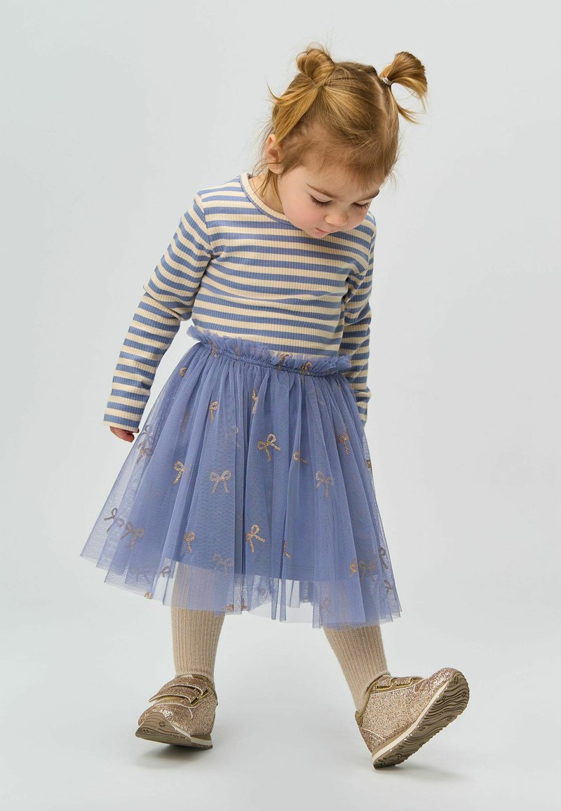 Gonna di tulle blu con motivi a fiocco dorato, abbinata a una maglietta a righe a maniche lunghe. Scarpe dorate glitterate e leggings beige completano l'outfit.