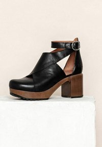 Zwarte leren enkelband schoen met een ronde neus en een uitgesneden design, voorzien van een bruine blokhak en een gestructureerde zool. Bevestiging met een gesp.