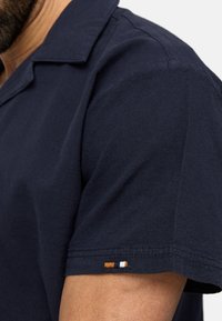 Camisa de manga corta de color azul marino con cuello, que presenta un tejido texturizado y un pequeño detalle bordado en la manga en color naranja, blanco y azul marino.