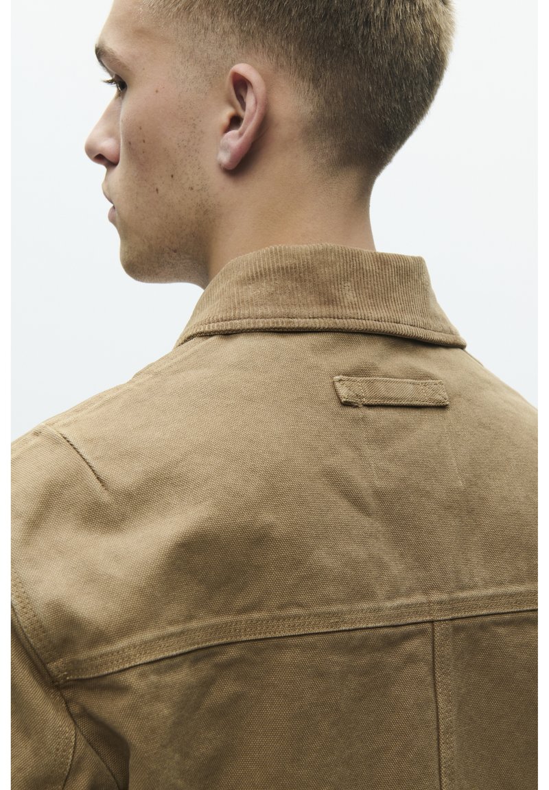 Veste en velours côtelé beige avec un col pointu, des détails de couture visibles et un patch carré au dos. Texture lisse, design décontracté.
