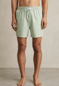 Shorts de bain vert clair avec une taille élastique et un cordon blanc. Fabriqués en tissu lisse, coupés juste au-dessus des genoux.