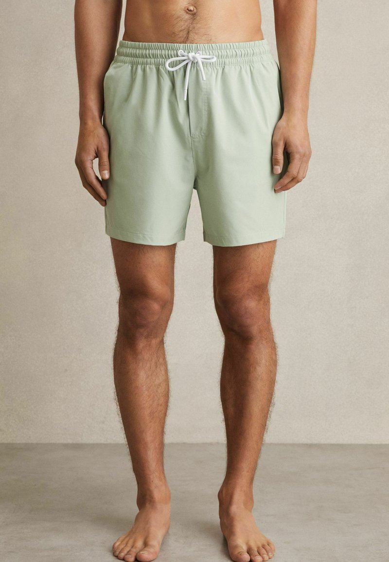 Shorts de bain vert clair avec une taille élastique et un cordon blanc. Fabriqués en tissu lisse, coupés juste au-dessus des genoux.