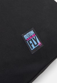 Zwarte stof tas met een gestructureerde buitenkant. Een rechthoekig label toont "BUTTON YOUR FLY" in blauwe en roze letters met een witte rand.