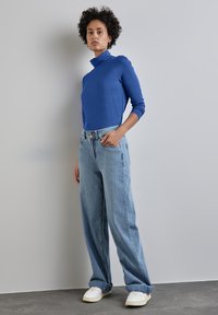 Blaues Rollkragenshirt mit langen Ärmeln, kombiniert mit hellblauen, weiten Jeans. Weiße Sneakers mit strukturierten Sohlen runden das Outfit ab.