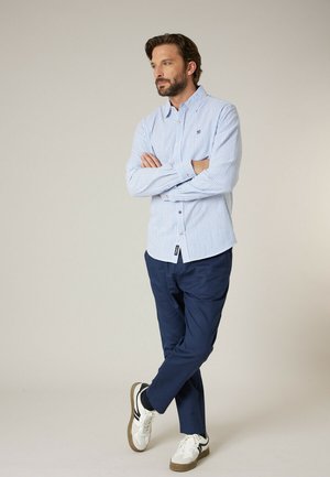 Man met baard staat met gekruiste armen, draagt een blauw gestreept shirt, marineblauwe broek en witte sneakers tegen een effen achtergrond.