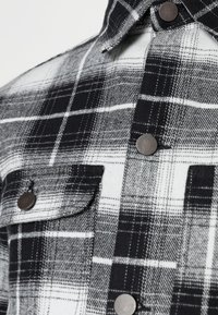 Svart och vit rutiga flanellskjortan med två bröstfickor, silverfärgade tryckknappar och en texturerad tyg med tydliga horisontella och vertikala linjer.