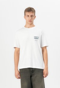 Ung mann med kort hår som har på seg en hvit t-skjorte med liten tekst på brystet og mørke, løstsittende jeans, stående mot en enkel hvit bakgrunn.