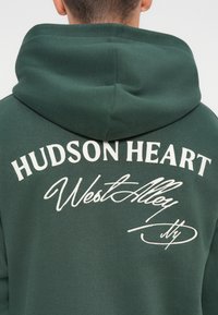 Zeleno kapuco z vrvico za nastavljanje, s belim besedilom: "HUDSON HEART" in "West Alley" v kontrastnih slogih na hrbtni strani.