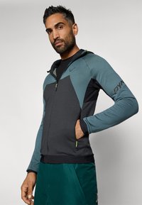 Férfi cipzáras kapucnis pulóver kevert anyagból, sötétszürke és petrolkék geometrikus mintával, fekete részletekkel és kapucnival.