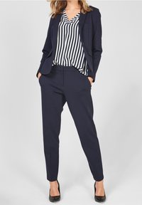 Completo con giacca blu navy e pantaloni, abbinato a una blusa a righe. La giacca presenta un design aderente e rever a booklet; i pantaloni hanno una vestibilità affusolata. Tacchi neri.