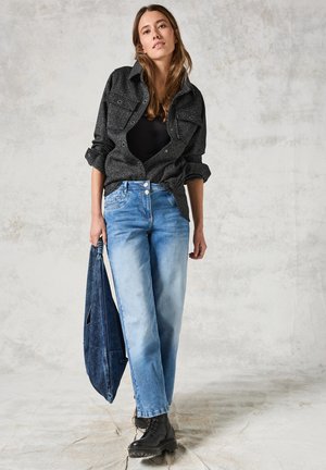Schwarze, strukturierte, übergroße Jacke mit zwei Brusttaschen, kombiniert mit hellblauen weiten Jeans und schwarzen Schnürstiefeln. Hält eine blaue Denim-Tasche.